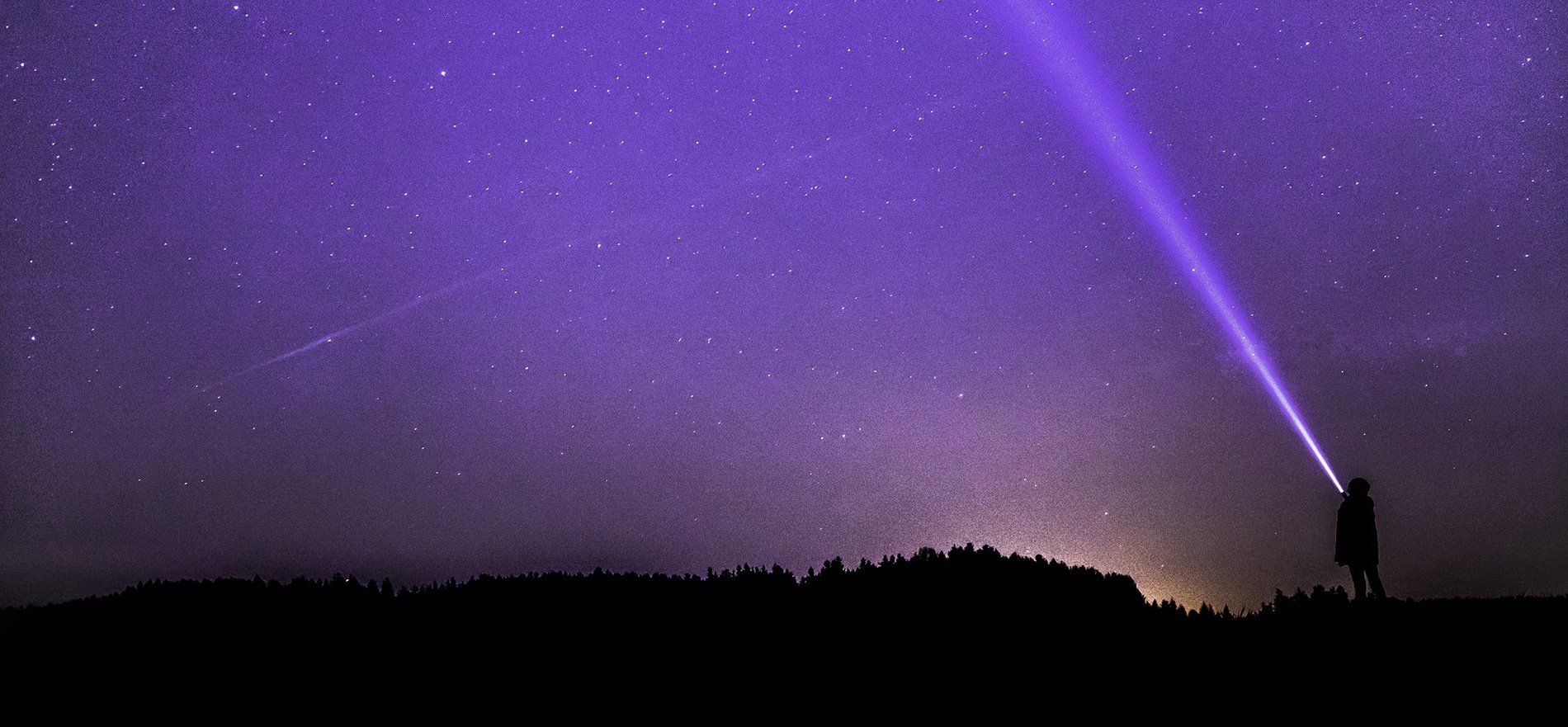 dark image, purple sunset with person on hill pointing a flashlight to the sky.