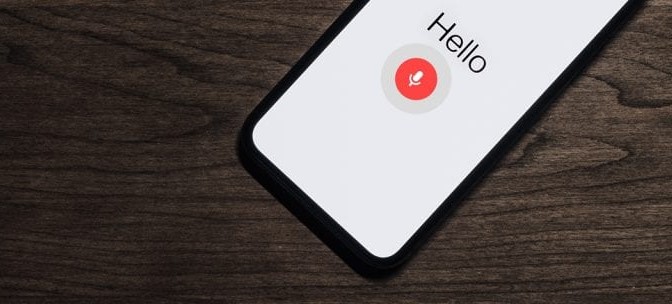 Cell phone on wood desk with the words "Hello" followed by a microphone button