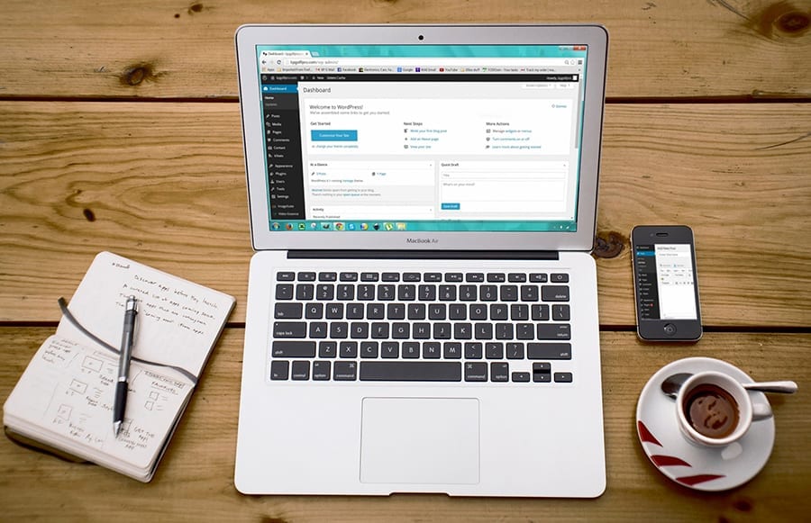 silver laptop with a phone, notebook and coffee mug sitting on a wooden table