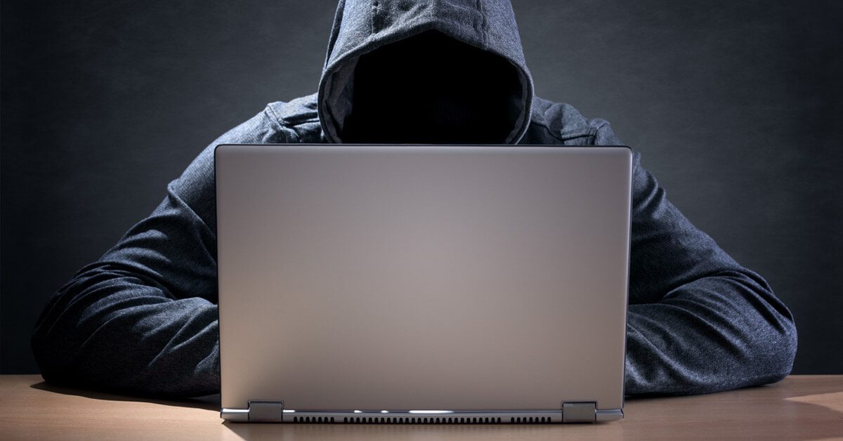 Laptop on table facing hooded man with face in the shadows
