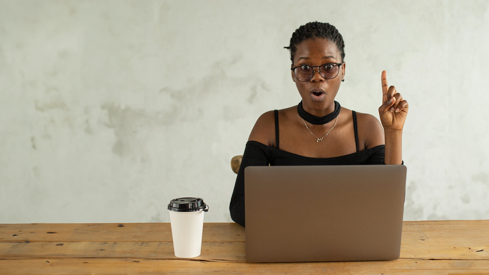 Dark skinned woman sitting at wood table with a silver computer and a coffee. Looking like she has a great idea with finger in the air pointing at the ceiling.